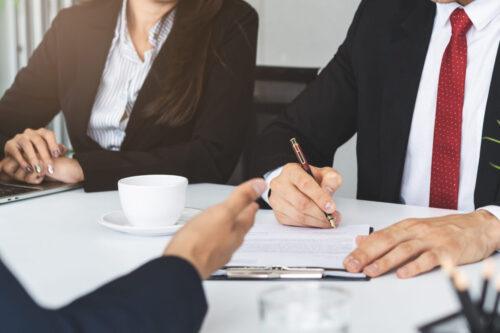 Two people in business attire sit at a table; one is signing a document while the other observes. A coffee cup and clipboard are on the table, suggesting a legal meeting about a medical malpractice case.