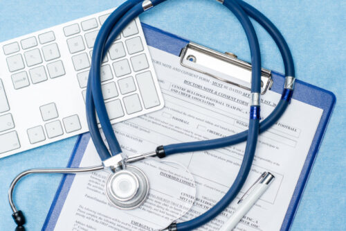 A stethoscope, pen, and eyeglasses rest on a medical consent form attached to a blue clipboard, with part of a white computer keyboard visible—a scene reflecting the importance of accuracy in medical records to help prevent malpractice.