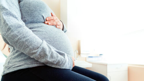 A pregnant person wearing a gray long-sleeve shirt sits in a bright medical office, possibly discussing concerns like hypoxic-ischemic encephalopathy, with one hand on their belly and the other on their lap. The background includes a counter and sink.